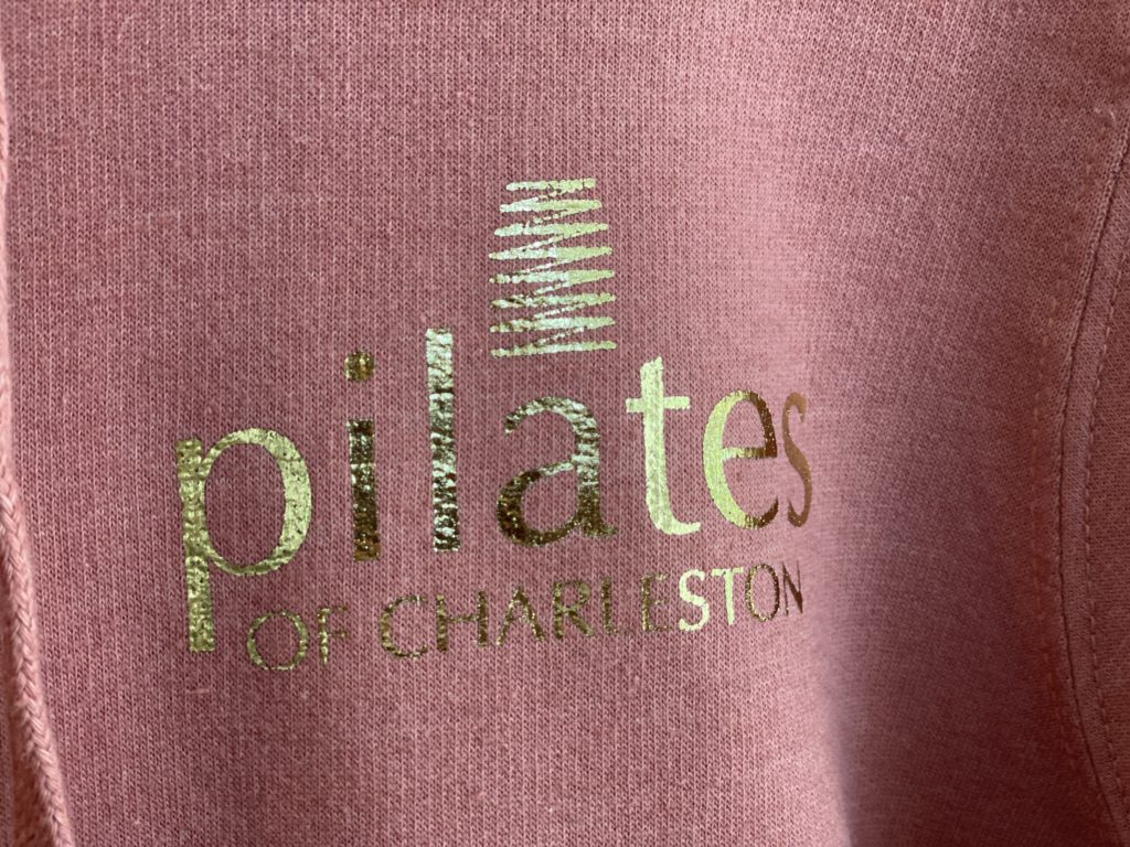 Logo of Pilates of Charleston on stylish pink activewear hoodie emphasizing Pilates classes and fitness in Charleston.