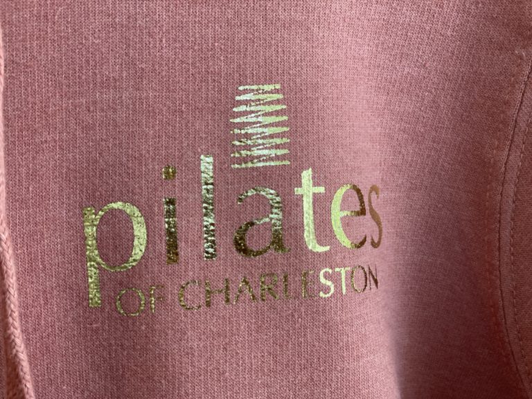 Logo of Pilates of Charleston on stylish pink activewear hoodie emphasizing Pilates classes and fitness in Charleston.